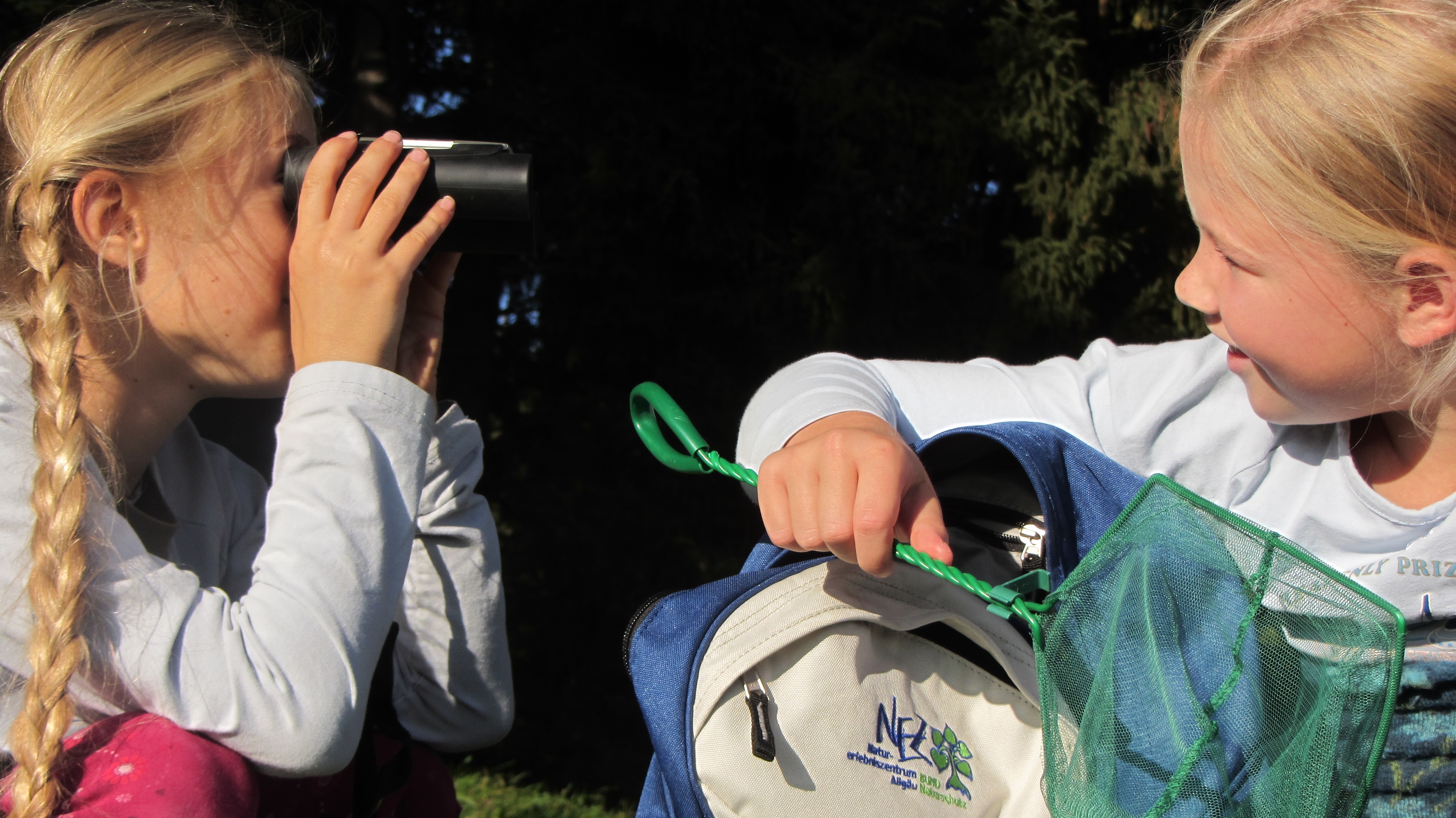 Ein Mädchen beobachtet ein anderes durch ein Fernglas, das zweite holt einen grünen Kescher aus einem Naturerlebnisrucksack